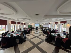 Zdjęcie sali z okrągłymi stolikami. Przy stolikach siedzą uczestnicy seminarium. Na wprost znajduje się ekran z wyświetloną prezentacją.